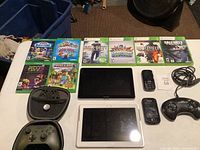 Overview photo of electronics and gaming items spread on table including game cases, Xbox One controller in case, tablets, phones, and Sega controller