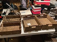 Three woven rectangular baskets with handles placed on a white table surrounded by various boxes and household items.