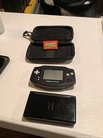 Nintendo DS Lite and Game Boy Advance handheld consoles with orange game cartridge in black protective case, and separate black carrying case on a table.