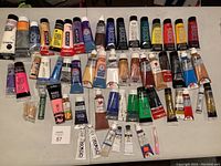 Full lot of assorted acrylic paint tubes arranged on table, showing brands and multiple colors.