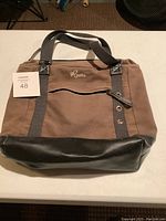 Full view of brown canvas handbag with black leather bottom and two black fabric straps, showing the Roots logo embroidered above the front zippered pocket.