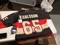 Back view of black Ottawa Senators hockey jersey showing Karlsson name and number 65 with red and white horizontal stripes and Reebok branding.