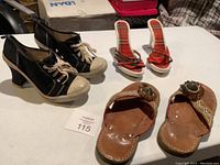 Lot of three pairs of women's shoes on a white table: black heeled sneakers, red plaid heeled sandals, and brown leather slip-on sandals with decoration.