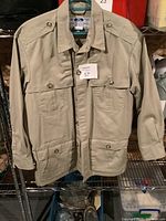 Full front view of khaki Tilley Endurables jacket hanging on blue hanger, with four buttoned front pockets and shoulder epaulets.