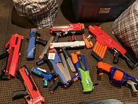 Photo showing various Nerf and water toy blasters arranged on carpet, mainly Nerf Rival models in red, blue, orange, white, and green colors.