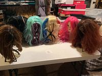 Six wigs on six black wig stands arranged on a table in various colors: light brown, mint green, platinum blonde with red and blue, bright pink, curly light brown, and black.