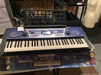 Front view of Yamaha keyboard with blue control panel, 61 keys with number stickers, large speakers, and music stand.