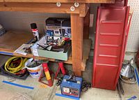 Photo showing jumper cables coiled in a bucket, tire chain, CenTech digital inspection camera in box, two red plastic car ramps, blue battery charger, two red hydraulic jacks, and spray can on floor under a wood bench.