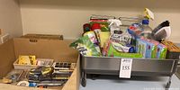 Metal dish rack loaded with cleaning products and a cardboard box with tools and hardware items next to it on a table
