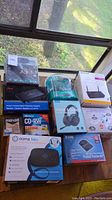 Photo of multiple boxed items including network switch, Ooma Telo phone, Netgear access point, Logitech headset, Coosii headphones, D-Link router, and blank media discs.