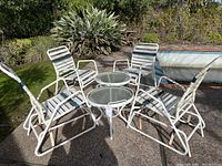 Four white vinyl and aluminum outdoor chairs placed around two round glass-top side tables outside on a patio, showing rust and weathered condition.