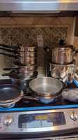 Stack of polished stainless steel pots with lids except one 10" two-handled pot without lid, all on stovetop.