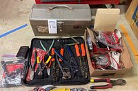 Full view of metal toolbox, black roll-up electrician tool case with red, orange, blue handled tools, and cardboard box with assorted cables and testing device