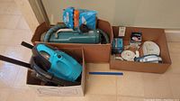 Boxes containing a vintage Electrolux canister vacuum, a small blue canister vacuum, white and blue handheld vacuums, and assorted vacuum parts