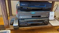 Stack of three video players on a low shelf: top Panasonic PV-8450 VHS player, middle Samsung DVD and VHS combo player, bottom Panasonic PV-9451 VHS player with remotes to the left
