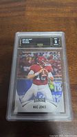 Front view of the Mac Jones rookie card graded GMA 9 in protective casing displayed on wooden surface.