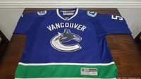 Full view of front of blue Vancouver Canucks hockey jersey in size XL with green and white stripe accents on bottom and sleeves, large team logo and 'Vancouver' text.