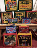 Photo showing 11 colorful collectible metal tins, mainly Crayola themed with crayons and vintage illustrations, alongside candy branded tins like Dubble Bubble, Tootsie Pops and Assorted Sours.