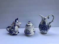 Photo showing 3 Delft Blue ceramic items including a cow creamer, lidded sugar jar, and floral pitcher against plain background