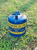 Blue galvanized steel oil can with large yellow label showing product and manufacturer details, placed on grass background.