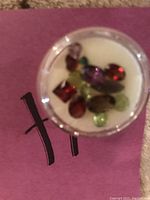 Close-up photo of multiple loose polished gemstones in red, purple, green, and brown tones contained inside a small clear round container.