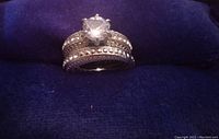 Photo of a silver tone women's ring with a large clear round gemstone centerpiece and two accompanying bands adorned with small clear rhinestones, displayed on a dark blue fabric background.