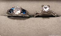 Close-up photo of two silver rings with clear stones, one having additional blue stones, placed in a grey jewelry display case.