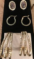 Photo showing three pairs of earrings: oval purple gemstone studs with rhinestone border, silver-tone braided hoops, and rhinestone embellished larger hoops.