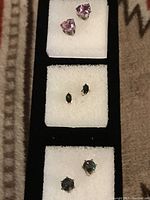 Three pairs of stud earrings displayed on white fabric pads, showing different colored gemstones in silver-tone metal settings.