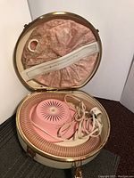 Open round cream-colored hard case showing vintage pink hair dryer with clear ribbed hose, power cord, and pink floral fabric interior lining.