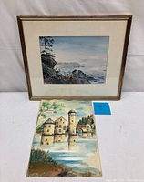 Photo of two watercolor paintings: one framed coastal landscape and one unframed water castle scene
