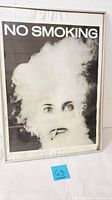 Framed black and white photograph poster titled 'No Smoking' depicting a face obscured by smoke with visible eyes and cigarette mouth, includes title text at top.