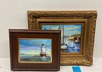 Two framed oil paintings; one of a lighthouse and the other of sailboats on water.