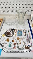 Photo of Fostoria glass bowl, textured glass vase, and a large assortment of colorful costume jewelry including various necklaces, earrings, and pins arranged on a white surface with a pegboard background.