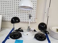 Photo showing two small black eyeball desk lamps and one large white adjustable eyeball desk lamp on a table against a pegboard wall.