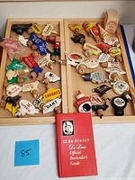 Top-down view of two wooden trays holding multiple vintage liquor bottle stoppers and a red hardcover bartender's guide book.