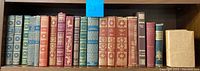 Full shelf view of 18 vintage leather-bound books in various colors with gold embossing.
