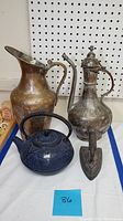 Photo showing a blue cast iron tea pot, antique sadiron, Turkish-style metal pitcher, and ornate metal coffee pot arranged on a flat surface