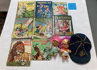 Photo of nine vintage children's storybooks, two troll dolls, and a Cub Scouts cap laid out on a white tablecloth.
