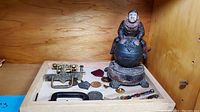 Photo showing vintage cast iron coin bank with seated figurine, brass drawer pulls, medals and pins, and pencils arranged in a wooden tray