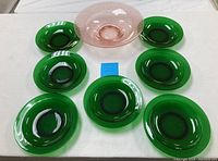 Top-down view showing seven emerald green salad plates surrounding one pink depression glass bowl with embossed pattern