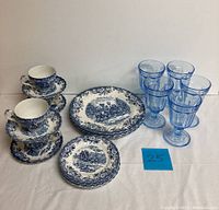 Photo showing the full lot with stacked Johnson Bros blue and white china cups, saucers, bread plates, dinner plates, and five electric blue stem glasses.