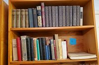 Wide angle photo capturing two wooden bookshelves filled with vintage books. Top shelf holds ornate hardcover books, bottom shelf features assorted vintage books.