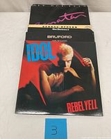 Stack of vinyl records with Billy Idol Rebel Yell in front, showing partial titles of other albums behind.