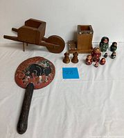 Photo shows all items in the lot: a small wooden wheelbarrow, a set of nine Russian nesting dolls, wooden container with floral decoration, Cape Cod salt and pepper shakers, and a Chinese animal design wall hanging fan.