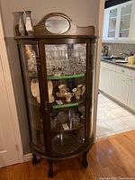Front view showing the curved glass door and shelves inside the oak curio cabinet filled with glassware and china items.