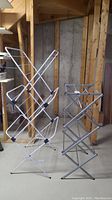 Two folding drying racks standing on a basement floor near unfinished wooden wall framing and concrete wall: one white multi-tier rack and one silver accordion-style rack.