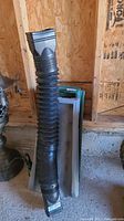 Two black flexible plastic downspout extenders standing upright against a wooden wall and concrete floor, showing ribbed accordion style design.
