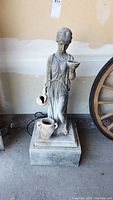 Front view of the classical style garden fountain statue depicting a woman holding a basin and jug, standing on a square base, showing natural weathering.