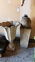 Two items side-by-side: a large concrete look planter with fluted flower edge design on a pedestal base and a tall pedestal with a spherical concrete look fountain top wrapped with black wire/cable.
